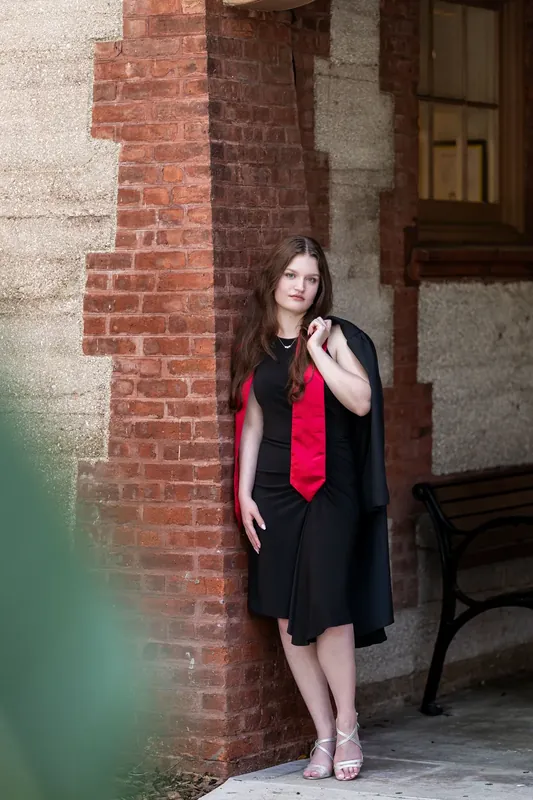 Olivia leans against red brick, holding her red honor cord and gazing thoughtfully to the side.