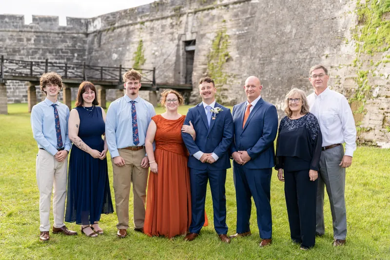 Nine people in navy, rust, and light blue formal wear stand shoulder-to-shoulder on grass with ivy-covered stone fortification behind them.