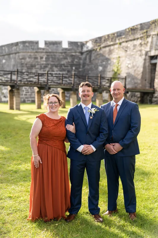 A person in a rust-colored gown stands with two people in navy suits on the fortress lawn, the ancient walls visible behind them.