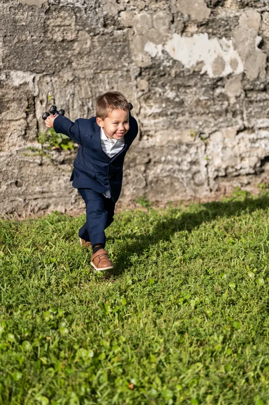 A young child in a dark suit runs across the grass in front of the fortress's weathered stone wall, looking back over their shoulder.