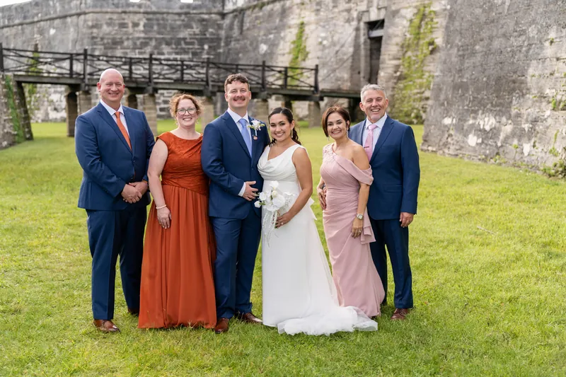 A person in white wedding attire stands center with family members in formal dress on both sides on the fortress grounds.