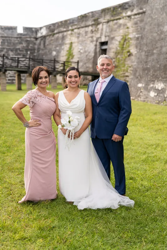 Patricia and Austin stand with a family member in formal attire on the grass in front of the historic stone walls, both smiling.