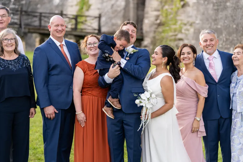 A group of roughly a dozen people in formal wear gathers on the lawn; one person holds a small child while others smile toward the camera.