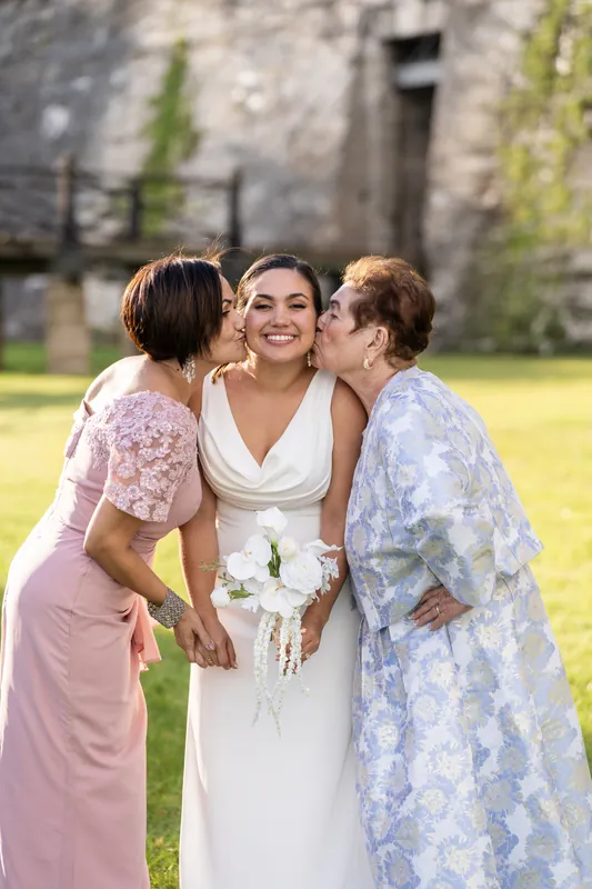 Person in a white wedding gown smiles as two guests in pink and blue dresses kiss their cheeks at the fort's grassy grounds.