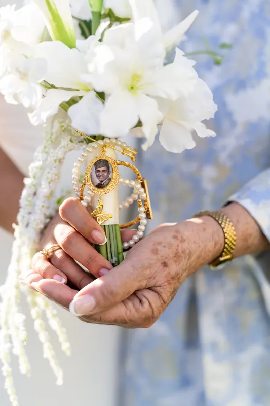 Hands holding a white bouquet with delicate flowers and a religious medallion against a blue sky backdrop.