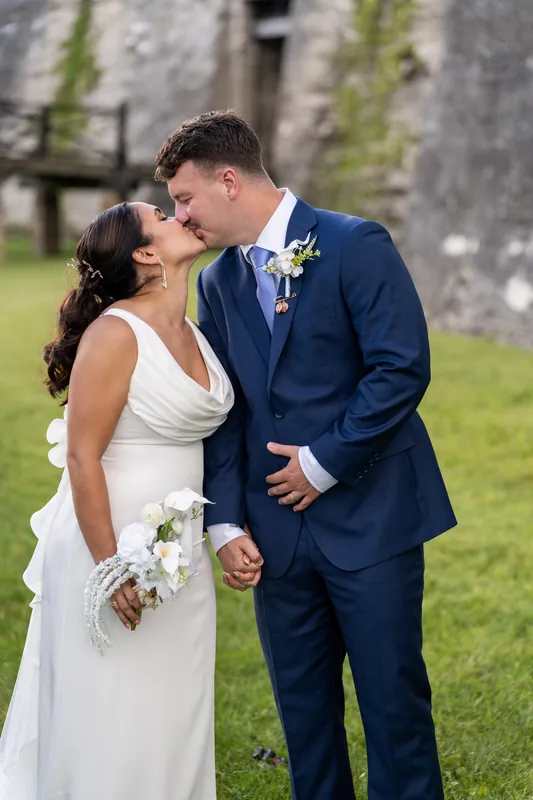 Two people in wedding clothes kiss while standing on the grass with the fort's weathered walls visible behind them.