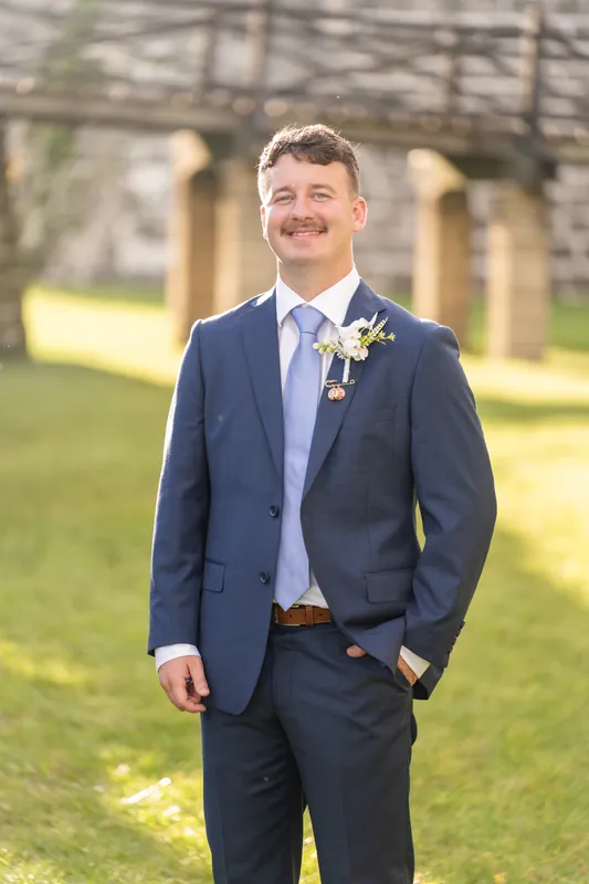 Person in navy suit and white shirt with boutonniere stands smiling on grass at Castillo de San Marcos, stone arches behind.