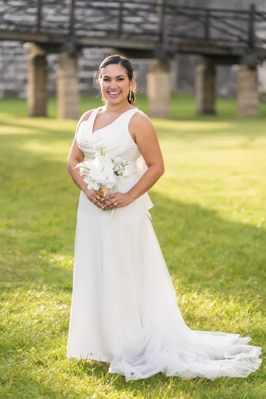 Person in sleeveless white gown holds white bouquet on the lawn at Castillo de San Marcos, stone walls visible behind.