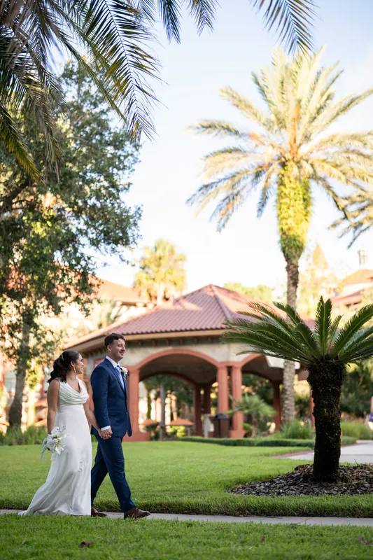 Two people walk across Flagler College grounds; one in white gown, one in navy suit, palms and terracotta building framing them.