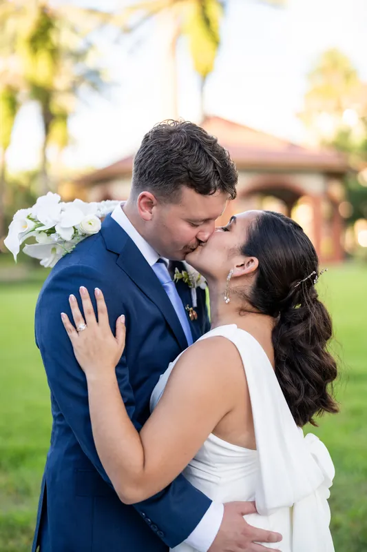 Patricia and Austin kiss on the lawn at Flagler College, her hand on his shoulder, white flowers and hedges around them.