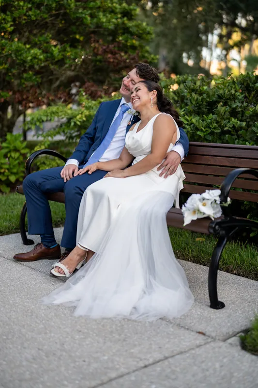 Two people sit on a wooden bench at Flagler College, embracing and smiling, surrounded by campus greenery.