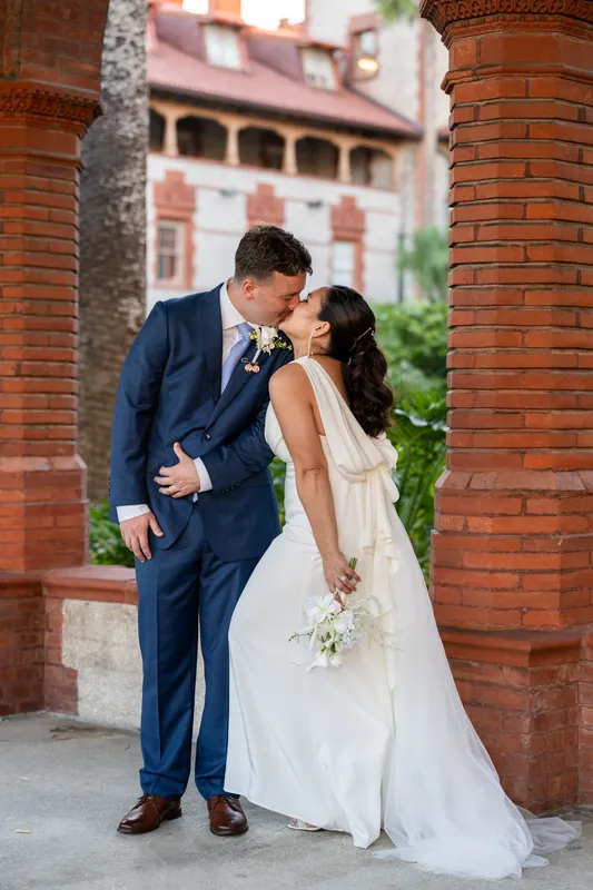 Patricia and Austin kiss beneath the brick archway at Flagler College, with colonial architecture and manicured grounds visible behind them.