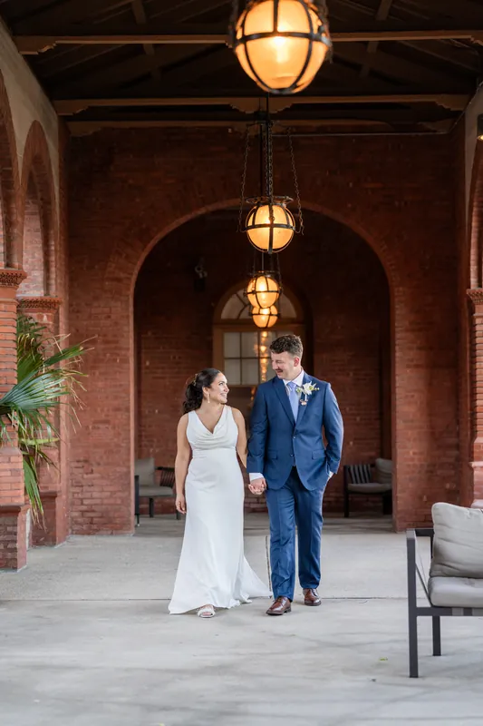 Patricia and Austin walk hand in hand beneath hanging lanterns in Flagler College's brick colonnade, moving toward an arched doorway.