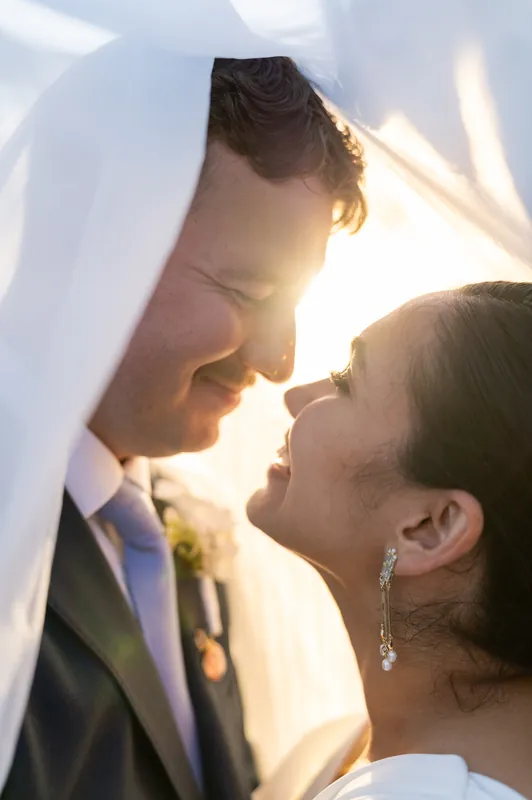 Two people in wedding attire share a close moment in bright sunlight, their faces nearly touching as golden light streams across their profiles.