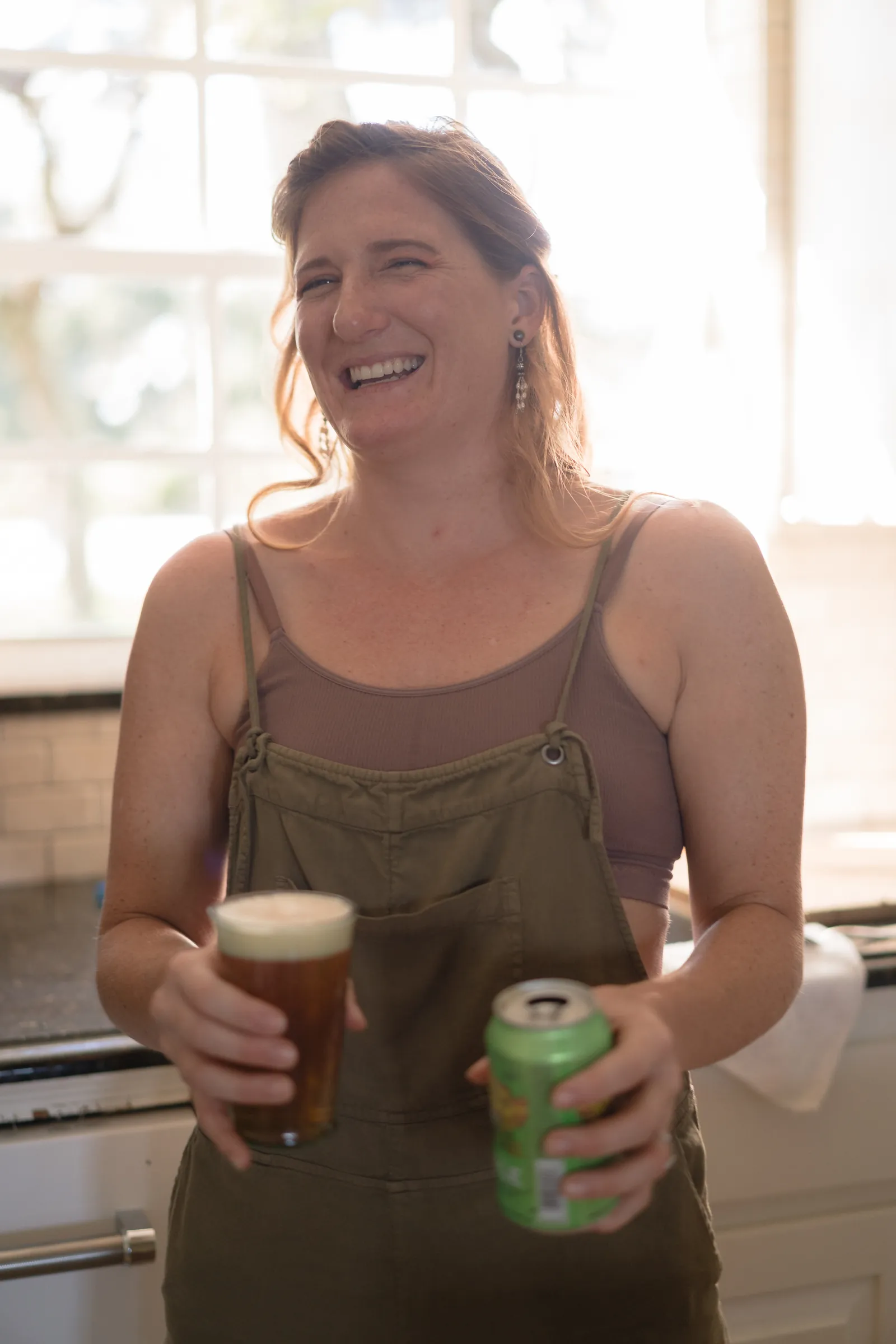 Bride in olive overalls laughing with beer during getting ready