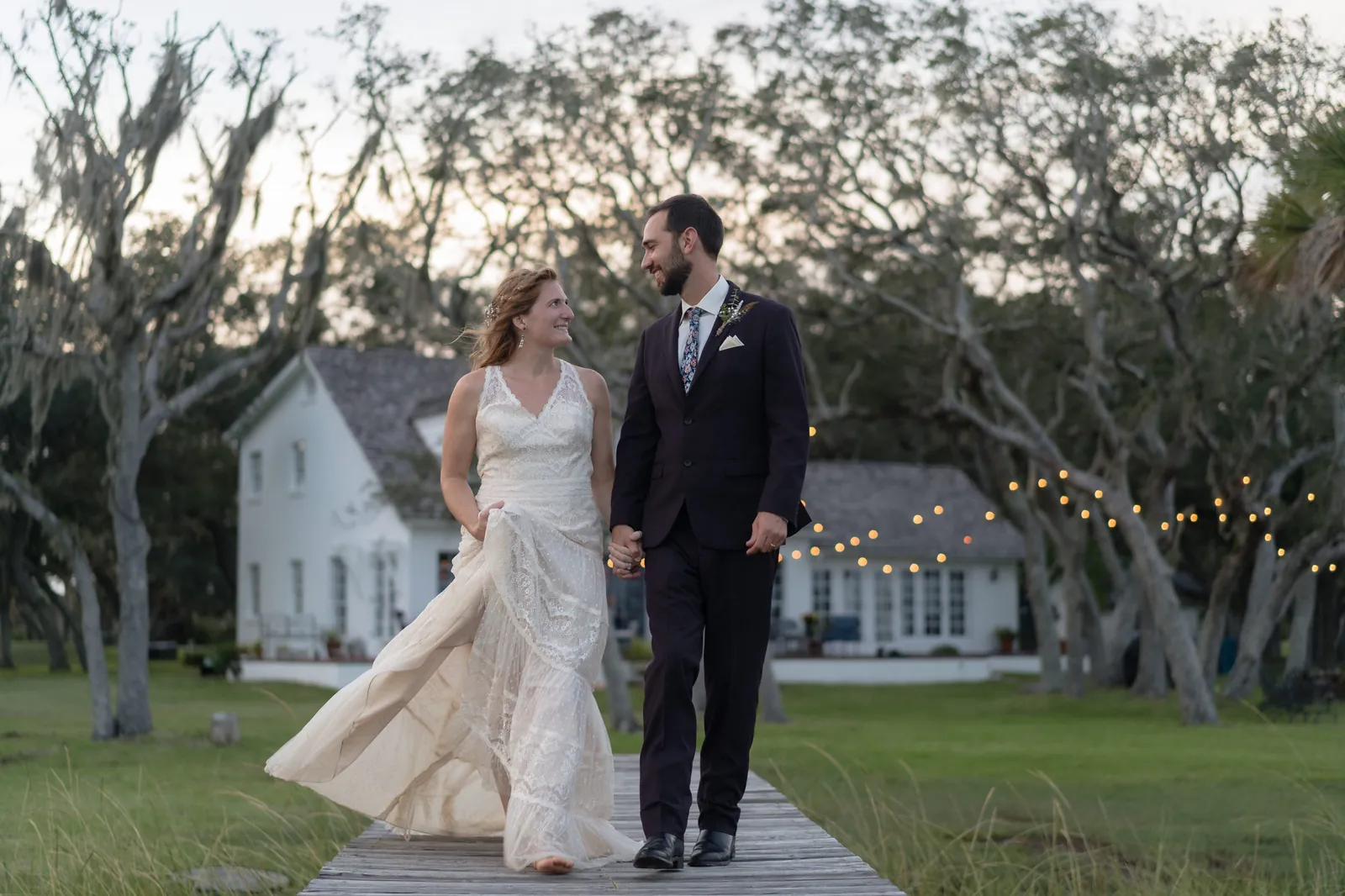 Couple walking on dock toward house with string lights at dusk