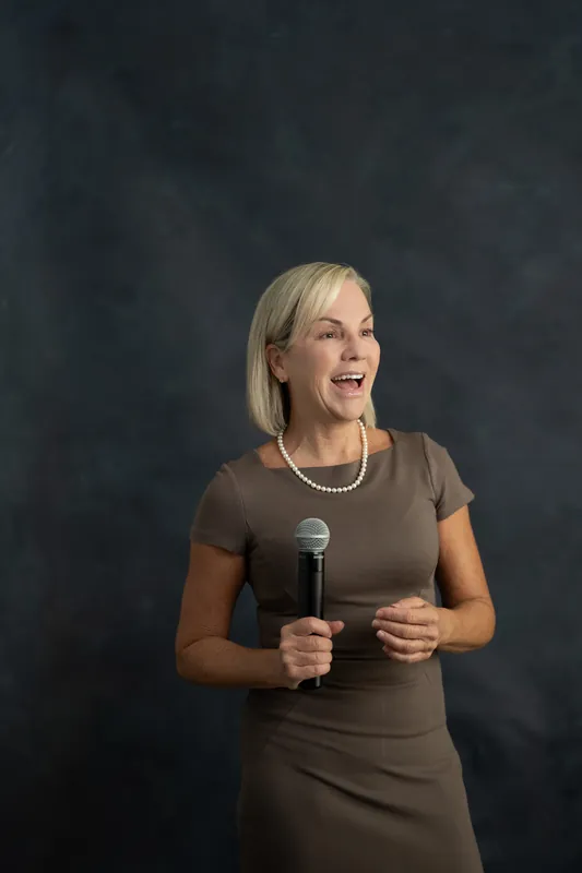 Paula Seymour speaks animatedly while holding a microphone at Casa Futrell in St. Augustine, wearing a brown fitted dress.