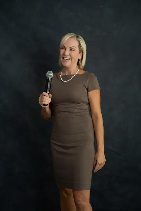 Paula Seymour stands poised with microphone in hand at Casa Futrell, smiling warmly in a brown short-sleeved dress.