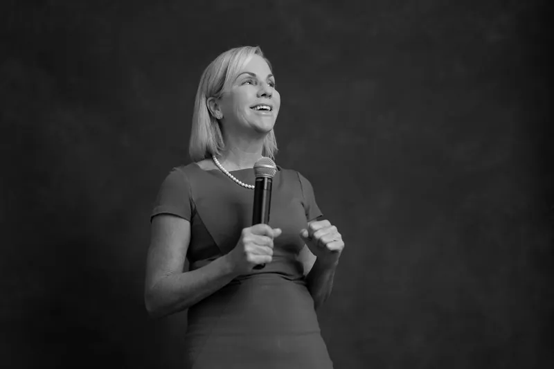 Paula Seymour gestures while speaking with a microphone, captured in black and white against a dark backdrop at Casa Futrell.