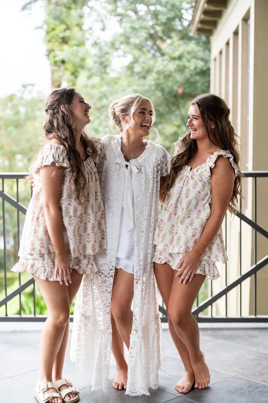 Three people in light lace dresses stand together on a covered porch overlooking green grounds at Crosswater Hall at Nocatee, Jacksonville.
