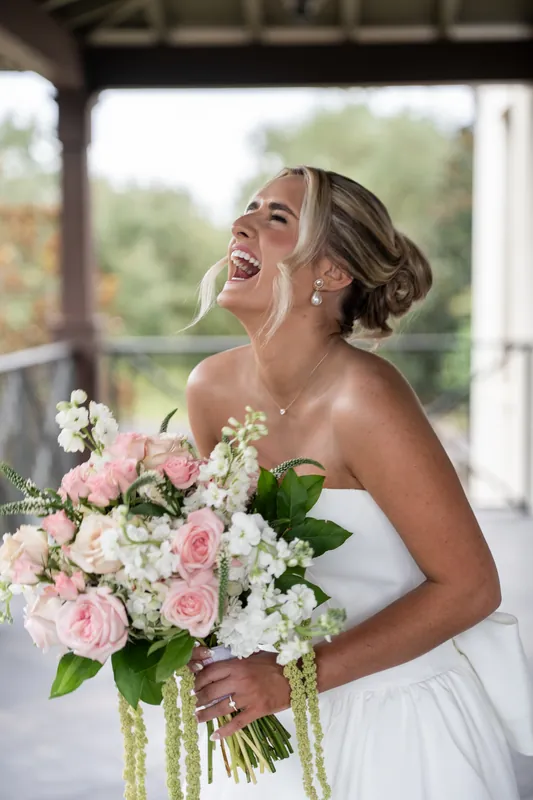 A person in a white strapless gown smiles while holding a pink and white bouquet on a covered porch at Crosswater Hall at Nocatee, Jacksonville.