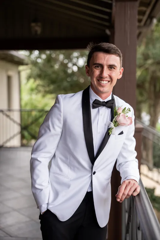 A person in a white dinner jacket and black bow tie leans against a railing, with soft evening light filtering through the portico behind them.