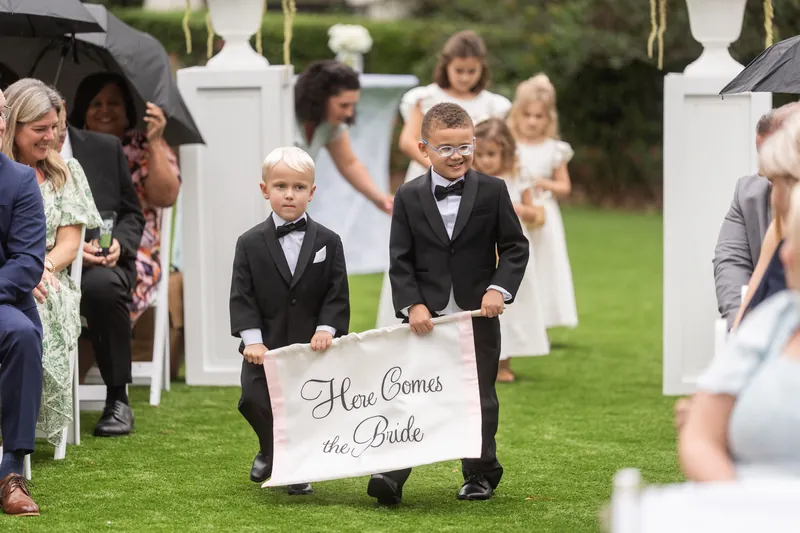 Two young children in formal wear hold a sign reading "Here Comes the Bride" on the lawn during the ceremony.