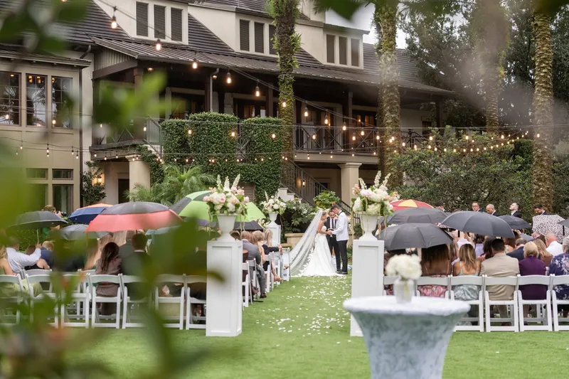 Outdoor reception setup with white folded chairs, tall floral arrangements, and guests beneath string lights and palm trees.