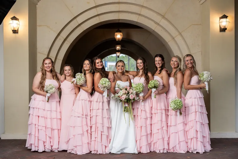 Bridal party of ten people in coordinating pink tiered gowns pose together under an arched stone entryway, holding white bouquets.