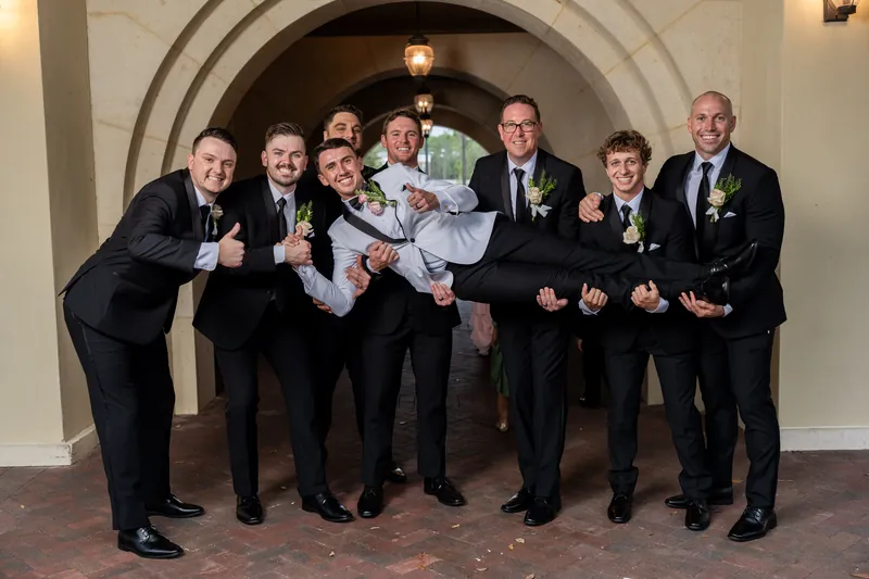 Eight people in dark suits with green boutonnieres hold a person in a white gown under an arched doorway at Crosswater Hall.