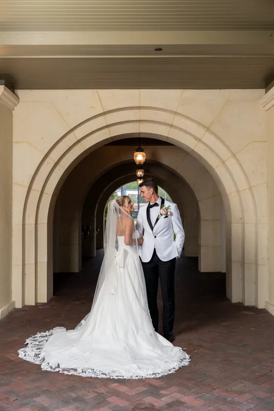 Payton & Tyler stand facing each other under a stone arch, one in a white gown with train and the other in a light blazer and dark pants.