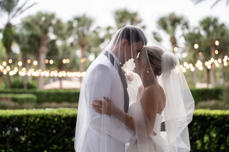 The couple kiss beneath a veil with strung lights and palm trees glowing softly in the background at Crosswater Hall.