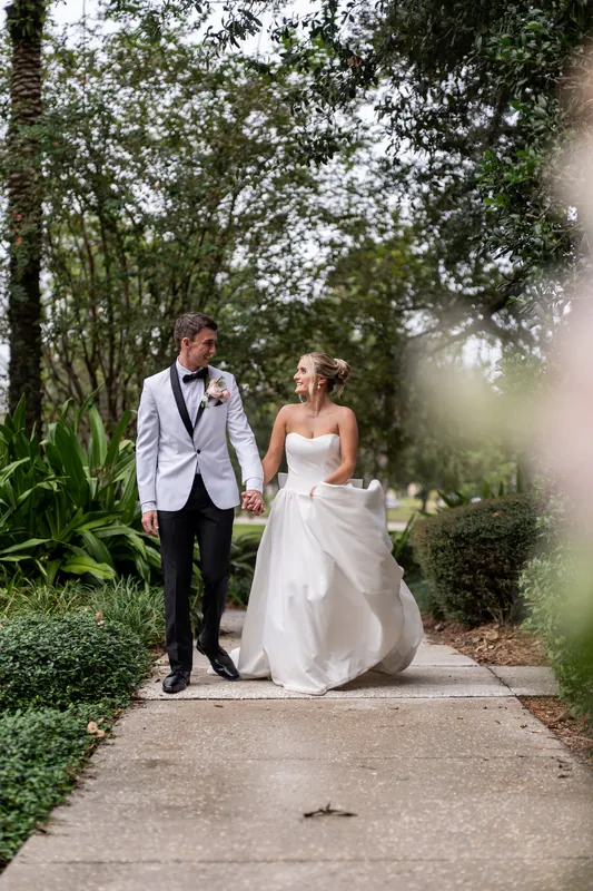 Two people walk hand-in-hand down a tree-lined garden path, one wearing a white gown and the other a light suit jacket.