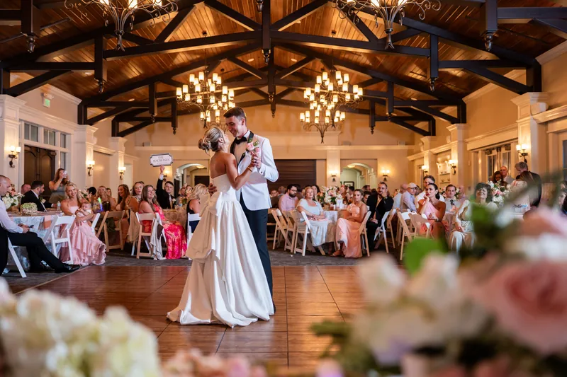 The couple dances in the center of a bright reception hall with wooden beams, chandeliers, and seated guests watching from tables.