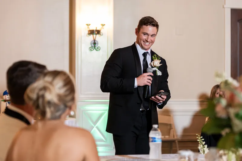 A person in a dark suit holds a microphone while giving a speech at a reception table, with a guest in the foreground.