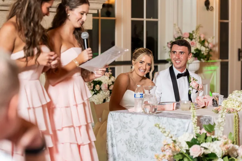 Two people in pink dresses speak at a microphone while Payton and Tyler sit at a table during their reception at Crosswater Hall.