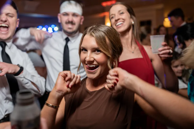 Wedding guests laugh and dance together on the dance floor, with a woman in a brown dress leading the celebration.