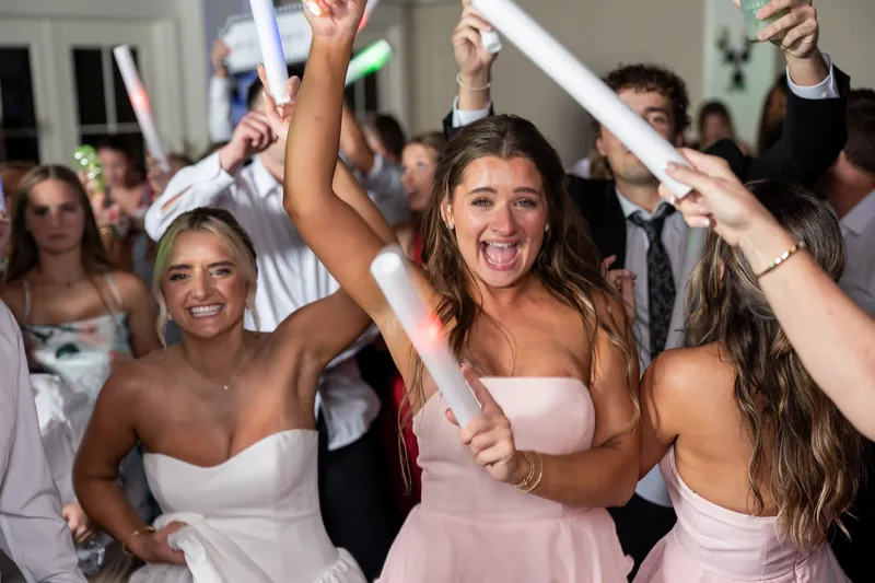 Women in white bridesmaid dresses raise their arms and cheer while holding ribbons during the reception.