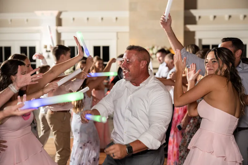A man in a light gray shirt and woman in a pink dress dance with guests waving glow sticks around them.