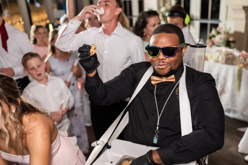 Man in sunglasses and black suit holds a microphone at the reception while guests dance behind him at Crosswater Hall.