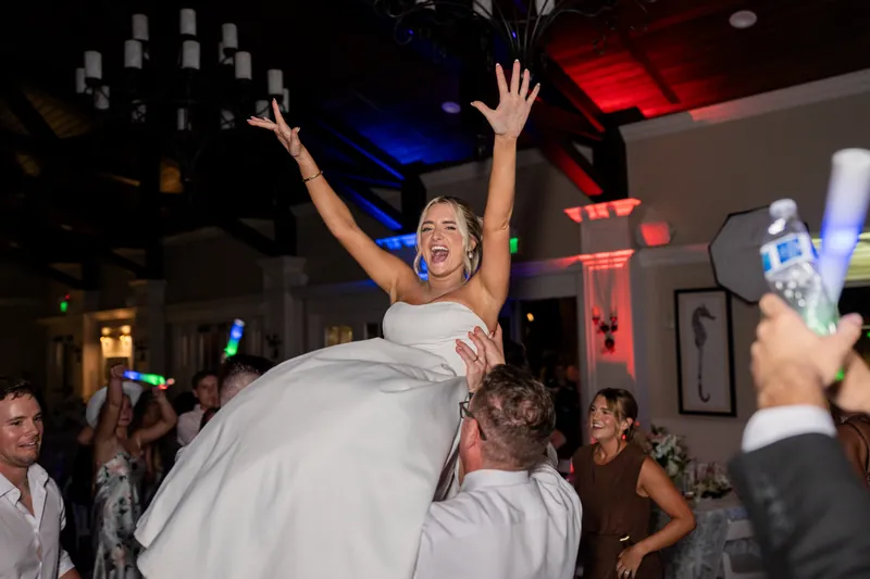 Person in white gown is lifted high by guests with arms raised in celebration during the reception.