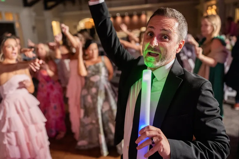 Man in dark suit with green neon accents raises his fist while entertaining guests on the dance floor.