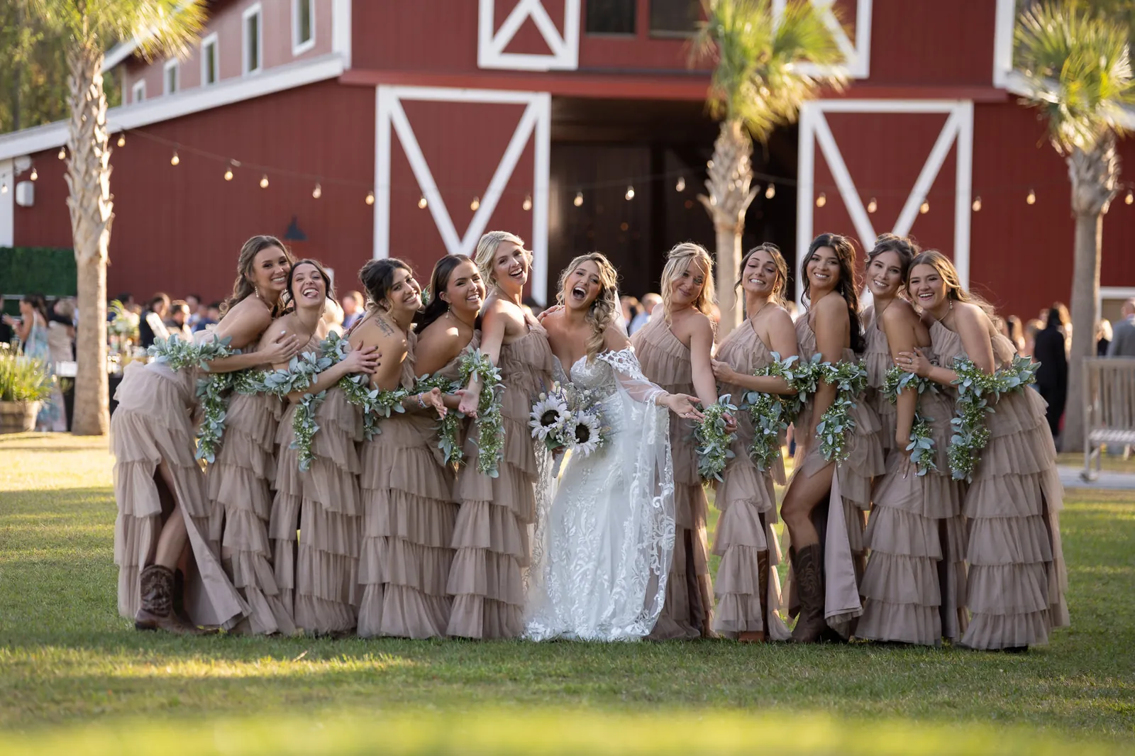 Bridesmaids laughing with bride in taupe dresses and greenery garlands at red barn Tringali Barn