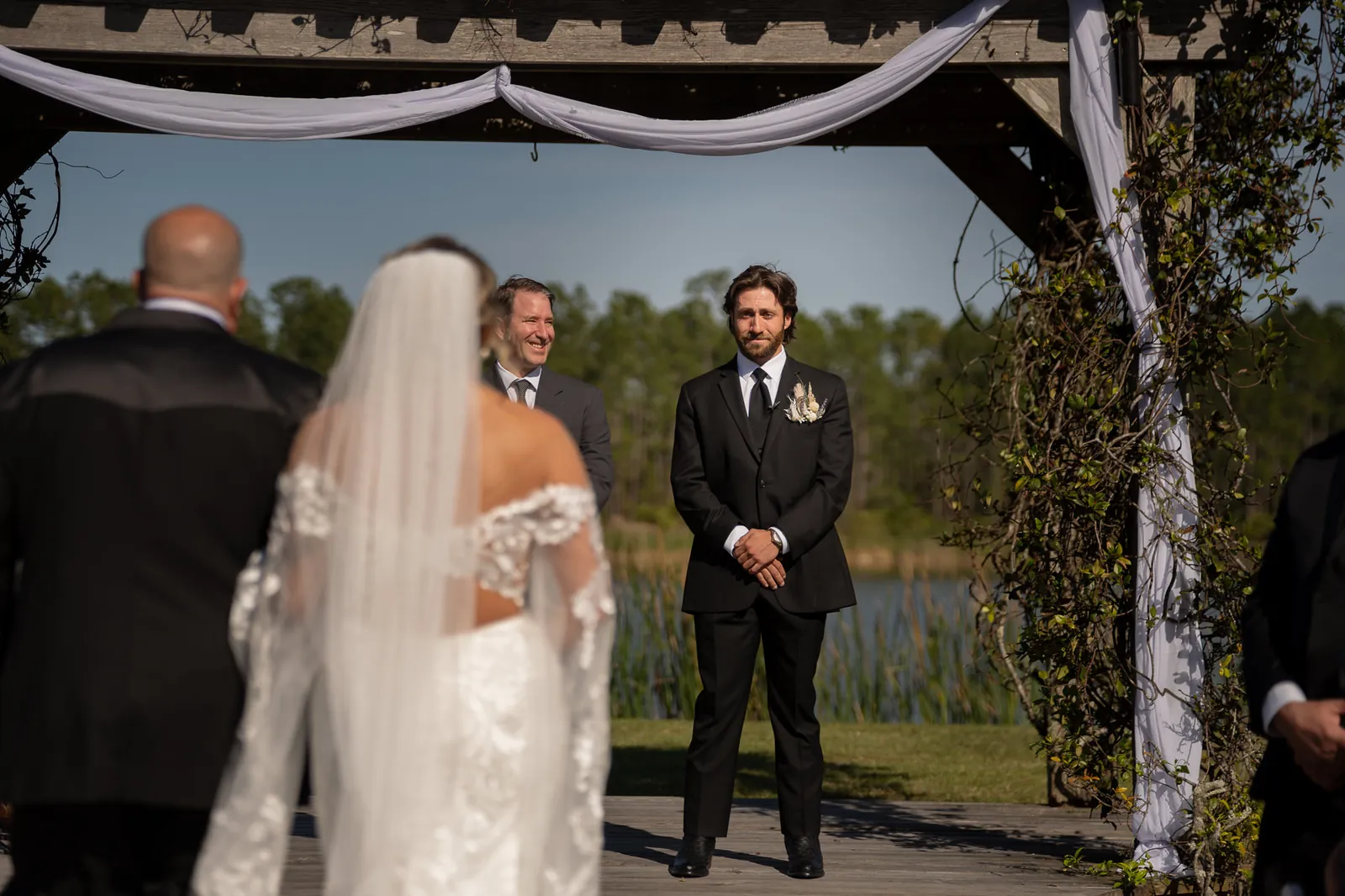 Groom Robert watching bride walk down aisle at lakeside pergola ceremony at Tringali Barn