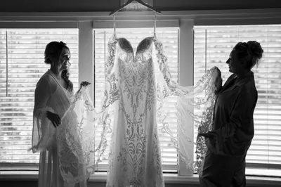 Bride's wedding dress displayed while two women admire intricate lace details indoors