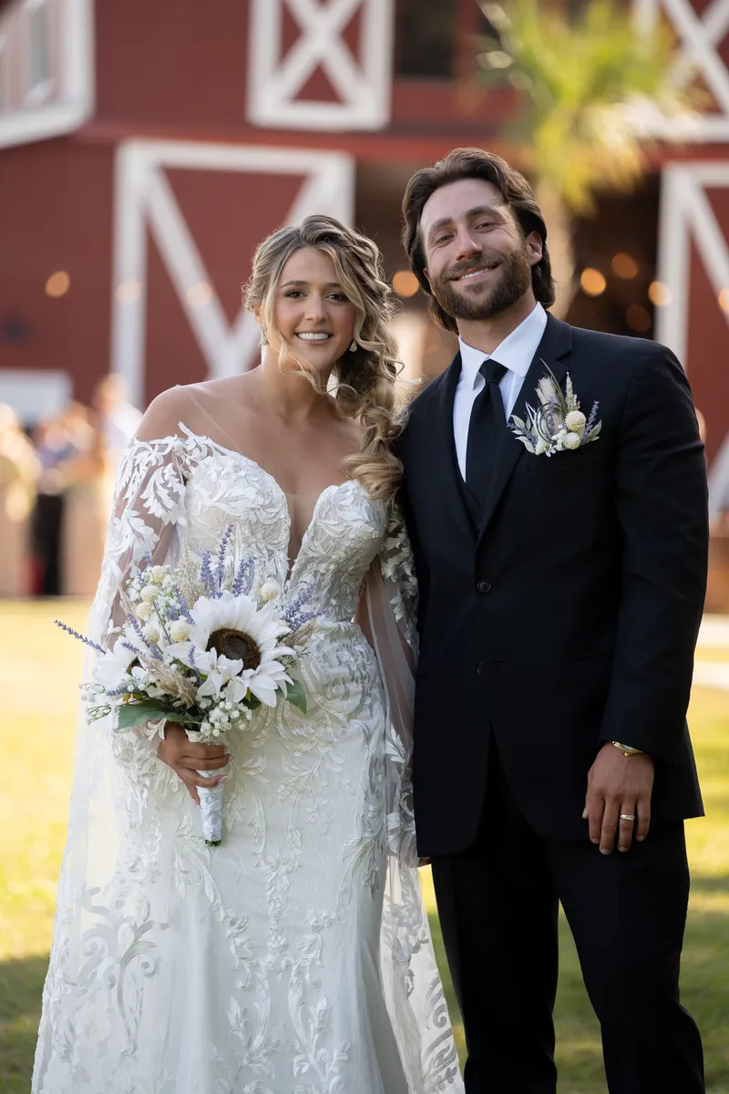 Riley and Robert stand on the lawn at dusk, Riley in a lace gown holding a white bouquet, Robert in a dark suit.