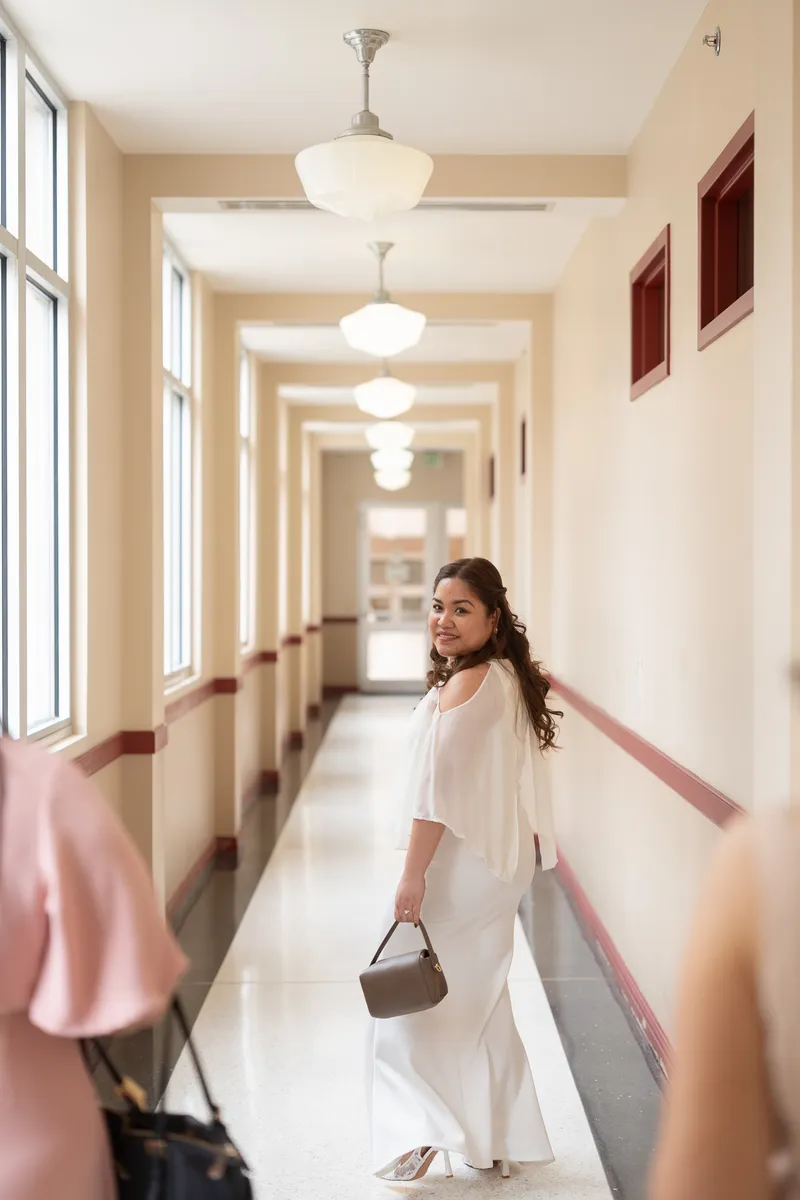 A bride in flowing white dress walks gracefully down an elegant hallway with vintage pendant lighting, looking back with a warm smile.