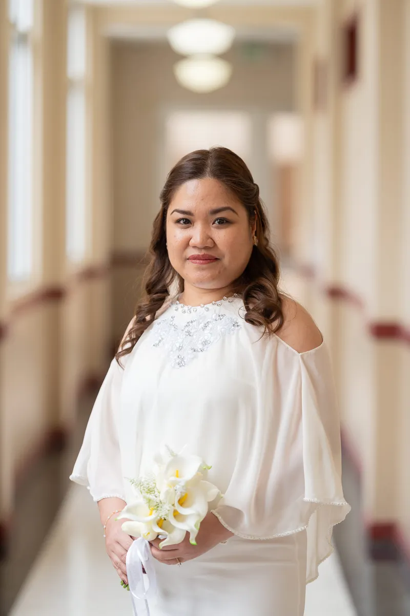 Vanessa poses elegantly in a white off-shoulder top with embellished neckline, holding a bouquet of white calla lilies in a bright hallway.