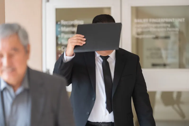 A man in a black suit and tie holds a folder or document up to shield his face while walking, with another person visible in the background.