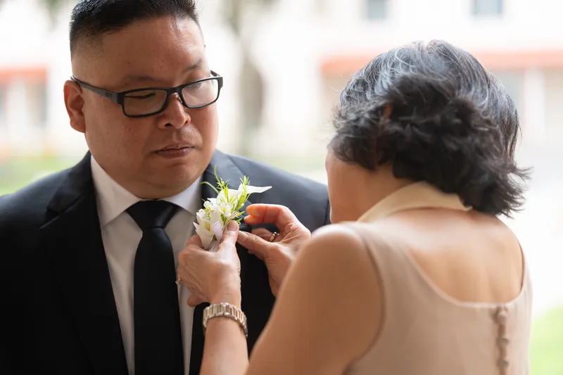 Vanessa lovingly pins a white floral boutonniere on Ronald's dark suit lapel in an intimate pre-ceremony moment.